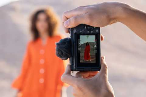 A pair of hands hold the Sony ZV-E10 II camera in front of a person with an orange dress.